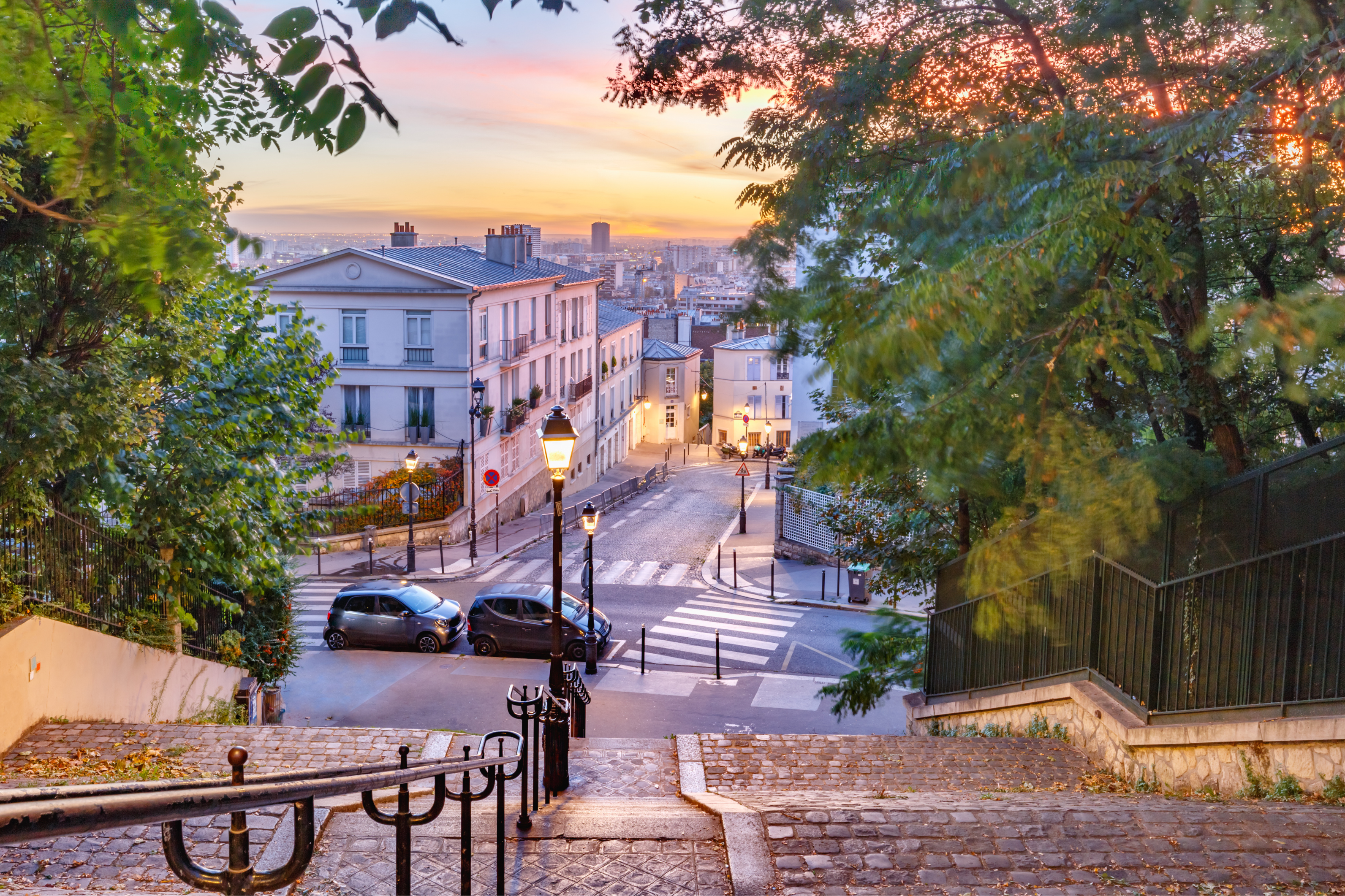 Une vue pittoresque d’un escalier pavé dans un quartier paisible de Paris (Montmartre) au coucher du soleil. La scène est encadrée par des arbres aux feuillages verts, des lampadaires classiques diffusant une lumière chaleureuse, et une rue calme bordée de voitures stationnées et de charmantes maisons résidentielles en arrière-plan. Le ciel se pare de nuances douces de rose et d’orange, créant une ambiance sereine et romantique.